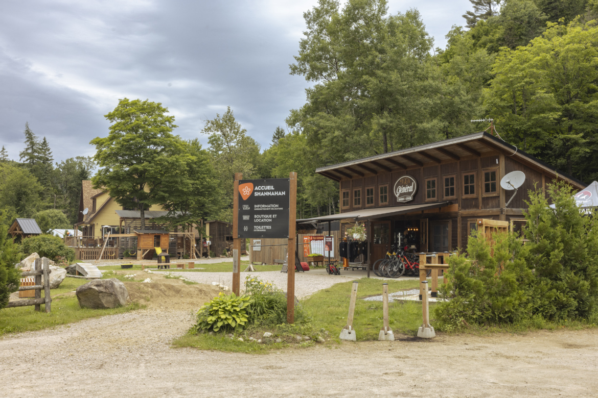 A rustic, wooden building serving as a welcome center and shop, surrounded by lush trees and a gravel path. A sign in front provides information about the facility, with various outdoor amenities visible, including bicycles for rent. The sky is partly cloudy, contributing to a scenic outdoor atmosphere.