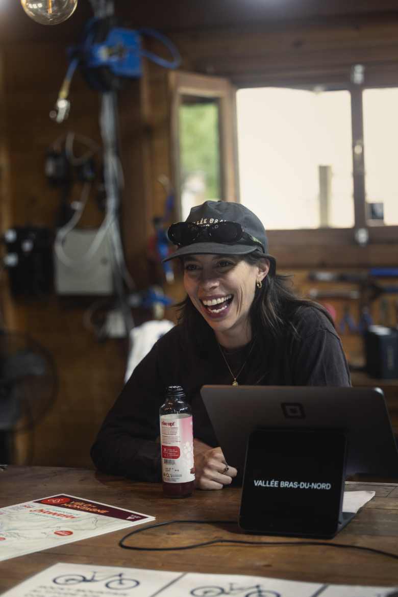A woman with long dark hair and sunglasses smiles joyfully while sitting at a wooden table. She is wearing a black cap and a black shirt, holding a drink in one hand. In front of her, there are maps and a tablet displaying the text "VALLÉE BRAS-DU-NORD" on the screen. The background features a rustic wooden interior with tools and equipment.