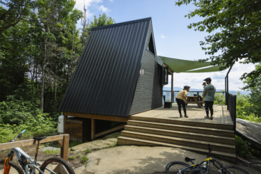 A modern A-frame cabin made of dark wood is set among lush green trees. Two people, wearing helmets, are engaged in conversation on the deck in front of the cabin, while a third person is visible in the background. The deck features a wooden table and a shaded area, with bicycles parked nearby. The landscape showcases a beautiful view of distant mountains under a partly cloudy sky. Empire 47 mountain bike trail.