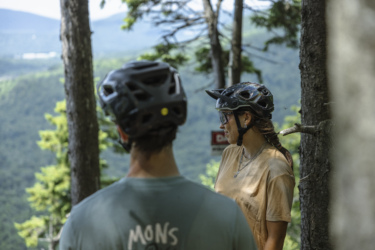 Two mountain bikers, one in the foreground wearing a helmet and a gray shirt, and another in the background wearing a helmet and a beige shirt, stand among trees. They are looking towards a scenic view of rolling green hills and distant mountains, with a sign visible in the background. The scene captures a moment of contemplation in a natural outdoor environment. Empire 47 mountain bike trail.