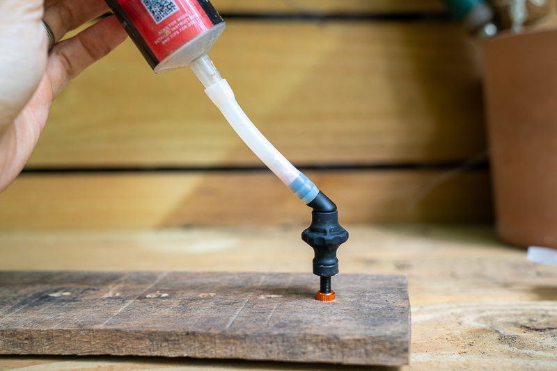 A person holding a bottle of adhesive above a wooden surface, with a flexible tube connected to a black dispensing nozzle positioned over an orange screw inserted into the wood. The background features wooden planks and a potted plant, creating a workshop environment.