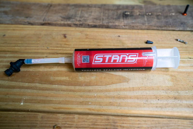 A tube of Stan's tubeless sealant injector is lying on a wooden surface. The injector features a black nozzle and a partially visible transparent chamber filled with sealant. Surrounding the injector are small bike components, indicating a maintenance or repair setting.