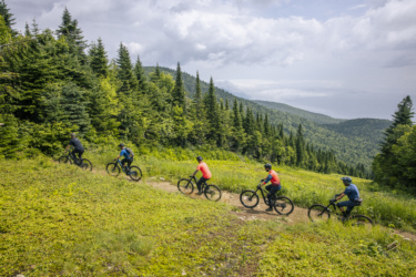 A group of five mountain bikers riding on a dirt trail surrounded by lush green trees and hills under a partly cloudy sky. The riders are dressed in colorful athletic attire, navigating through a scenic outdoor landscape. Le Massif de Charlevoix mountain bike trail.