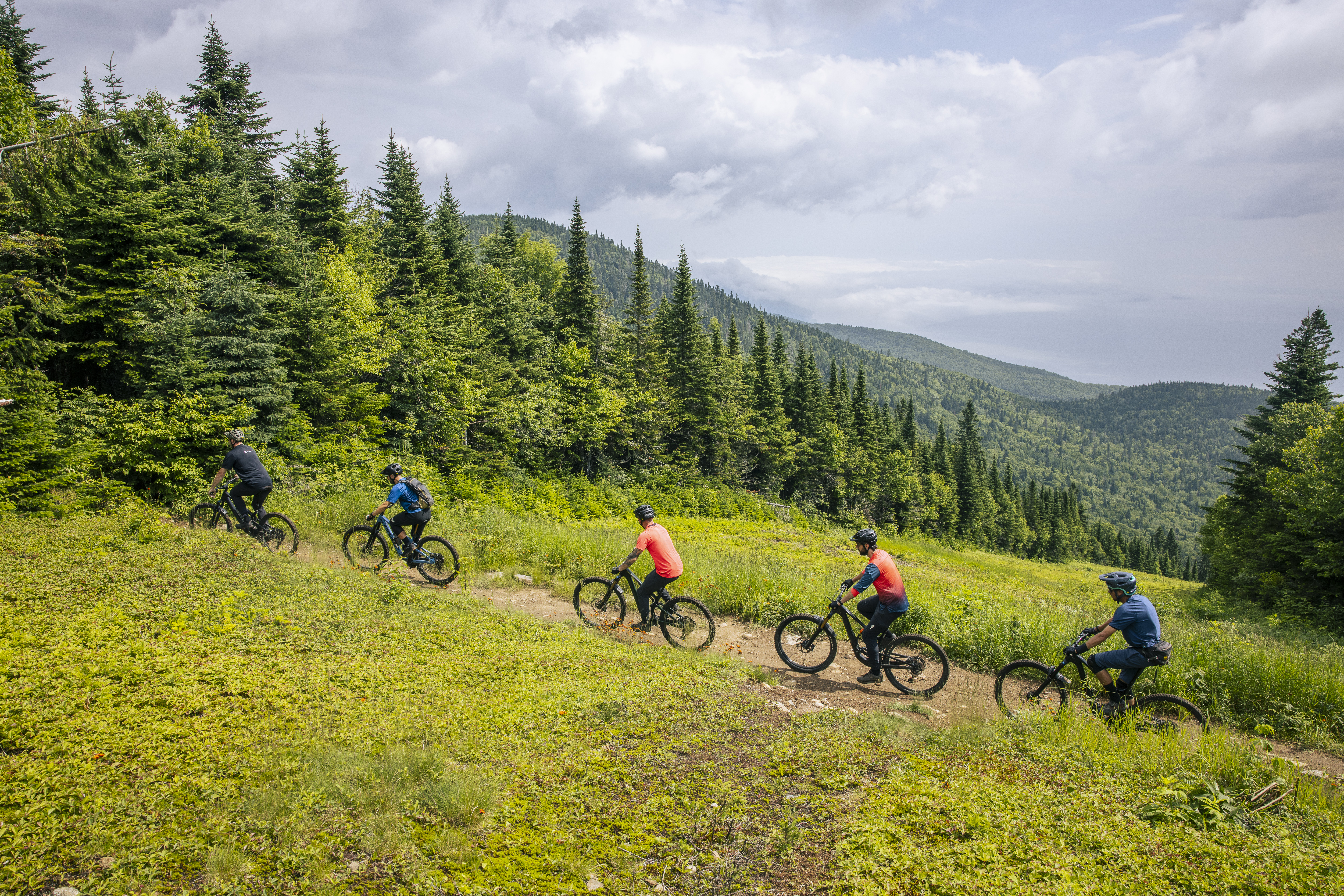 A group of five mountain bikers riding on a dirt trail surrounded by lush green trees and hills under a partly cloudy sky. The riders are dressed in colorful athletic attire, navigating through a scenic outdoor landscape. Le Massif de Charlevoix mountain bike trail.