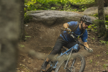A mountain biker navigating a winding trail through a densely wooded area, with rocks and dirt surrounding the path. The cyclist is wearing a helmet and protective gear, showcasing a focused expression as they lean into the turn on their blue bike. Lush greenery fills the background, adding to the adventurous atmosphere of the scene. Le Massif de Charlevoix mountain bike trail.
