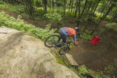 A mountain biker in a colorful jersey rides down a rocky ledge in a forested area. The cyclist is leaning forward, navigating the steep terrain. Lush greenery surrounds the rocky section, and a wooden ramp is visible in the background. Le Massif de Charlevoix mountain bike trail.