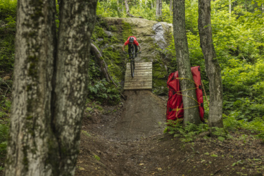 A mountain biker in a red shirt jumps off a wooden ramp in a lush green forest, surrounded by trees and rocky terrain. A red bag is positioned next to the ramp. Le Massif de Charlevoix mountain bike trail.