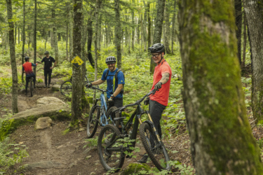 A group of mountain bikers rests on a trail in a lush, green forest. Two riders, one in a blue shirt and the other in a red shirt, smile as they pose with their bikes. In the background, two additional riders are visible, one interacting with the other. A yellow caution sign indicating a winding trail is seen on a nearby tree. The scene captures a moment of camaraderie and adventure in nature.