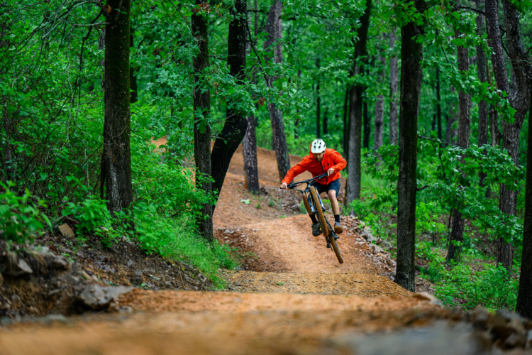 A mountain biker in an orange jacket and white helmet jumps off a dirt trail in a lush green forest. The scene captures the athlete mid-air, surrounded by tall trees and vibrant foliage.
