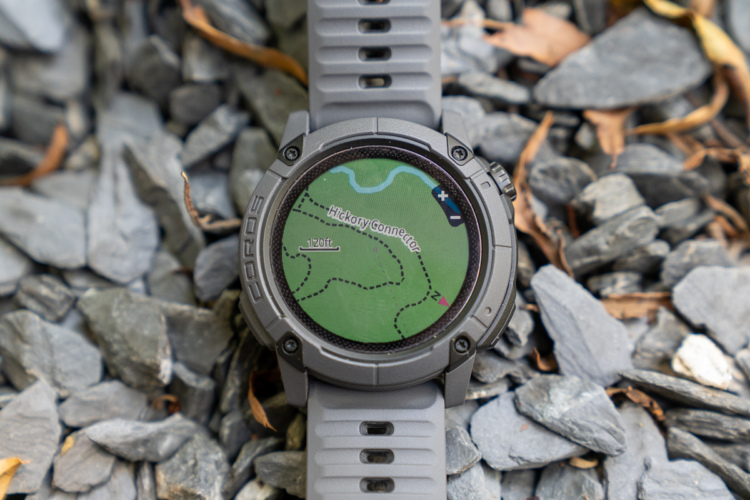 A gray GPS smartwatch displaying a topographical map with a highlighted hiking trail labeled "Hickory Connector." The watch is resting on a bed of gray gravel and small rocks, surrounded by a few dried leaves.