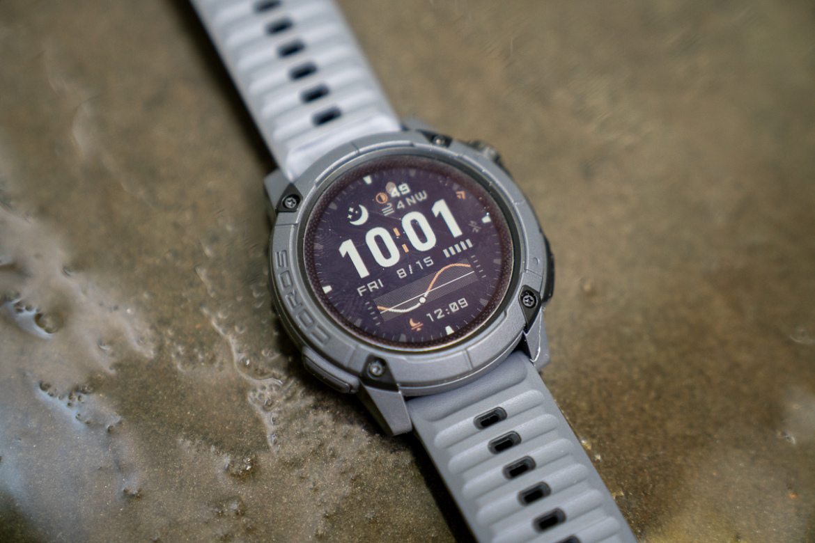A gray and black digital sports watch resting on a wet surface showing the time as 10:01, date as Friday 8/15, and an illuminated display featuring various icons and metrics. The watch has a silicone strap with ventilation holes, emphasizing its sporty design.