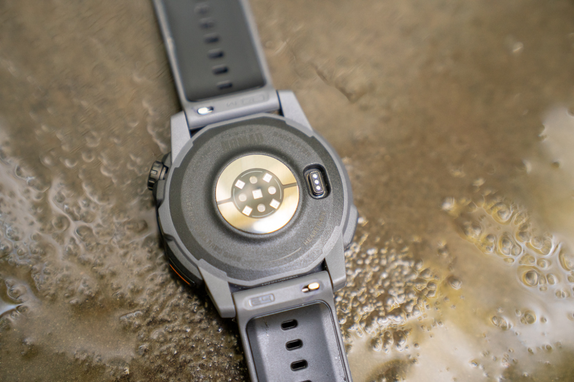 A close-up view of the back of a gray smartwatch, showcasing its sensor and charging ports. The watch is resting on a wet surface with droplets of water, highlighting its waterproof design.