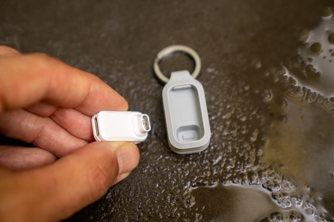 A close-up image of a person's hand holding a small white device with a charging port, next to a gray protective case with a keyring attachment. The background features a dark, textured surface with droplets of water.