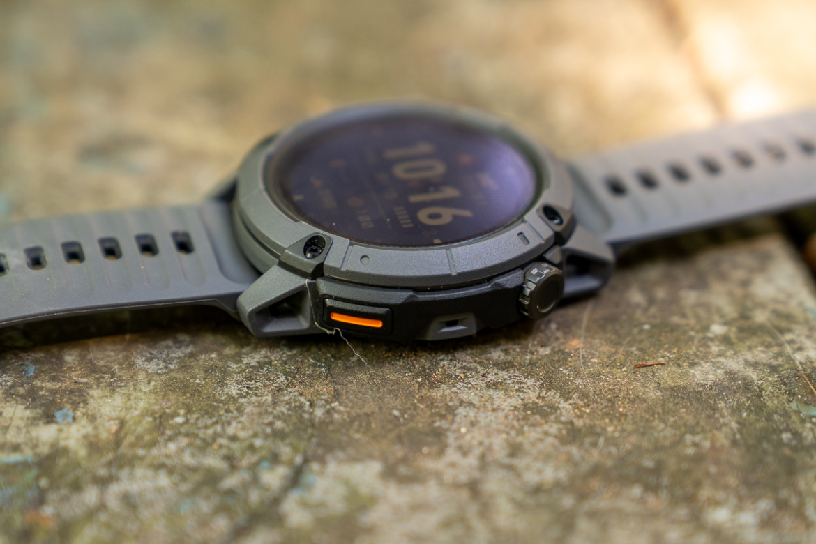 A close-up view of a rugged outdoor watch with a grey strap and an illuminated display showing the time and other functions, resting on a textured surface. The watch features a prominent knob and an orange accent on the side.