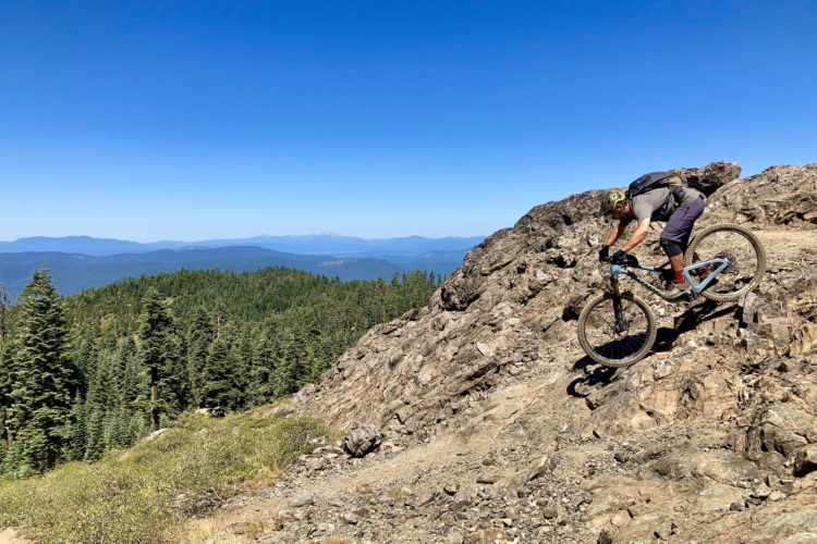 A mountain biker navigating a rocky trail surrounded by pine trees and a scenic landscape under a clear blue sky.
