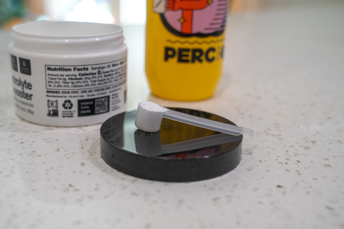 A close-up image of a measuring scoop with white powder positioned on a round black surface. In the background, there is a white container labeled "Electrolyte Booster" showing its nutrition facts, and a yellow beverage container partially visible with the word "PERC" on it. The image is set on a light-colored countertop.
