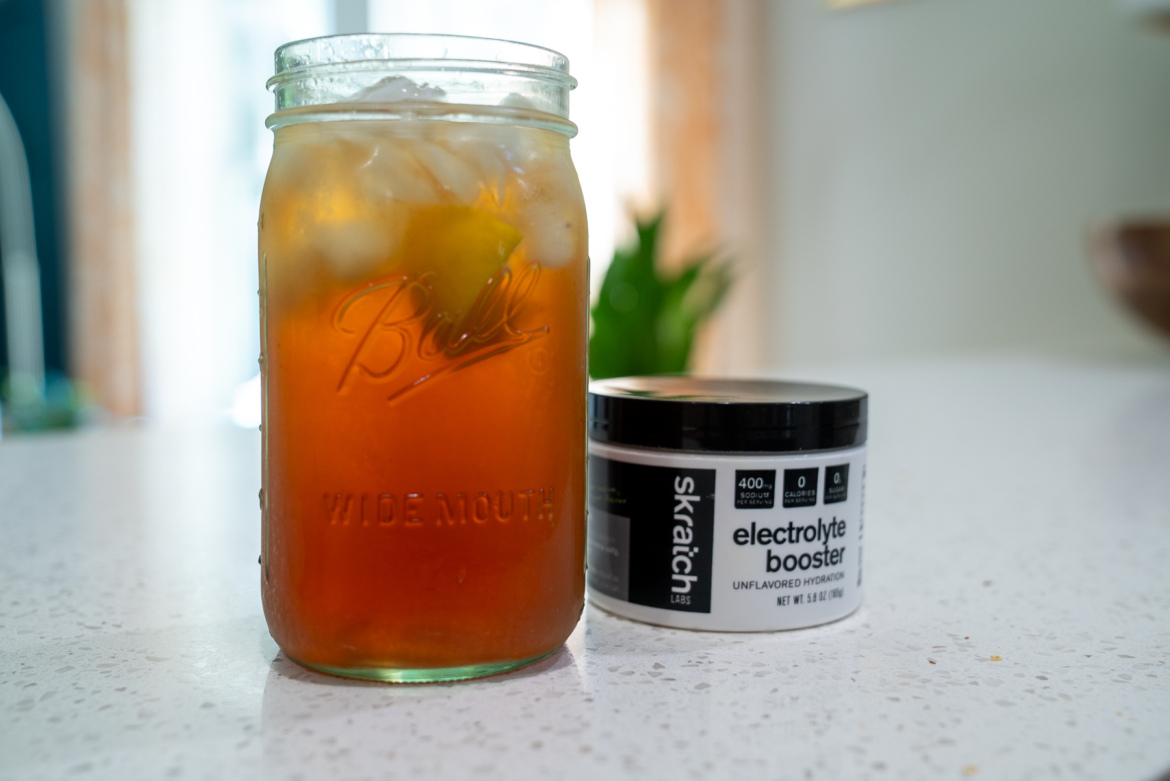 A glass jar filled with iced tea, ice cubes, and a lemon slice sits on a kitchen countertop next to a black container of Skratch Labs electrolyte booster, which is labeled as "unflavored hydration." In the background, there are hints of greenery and natural light.