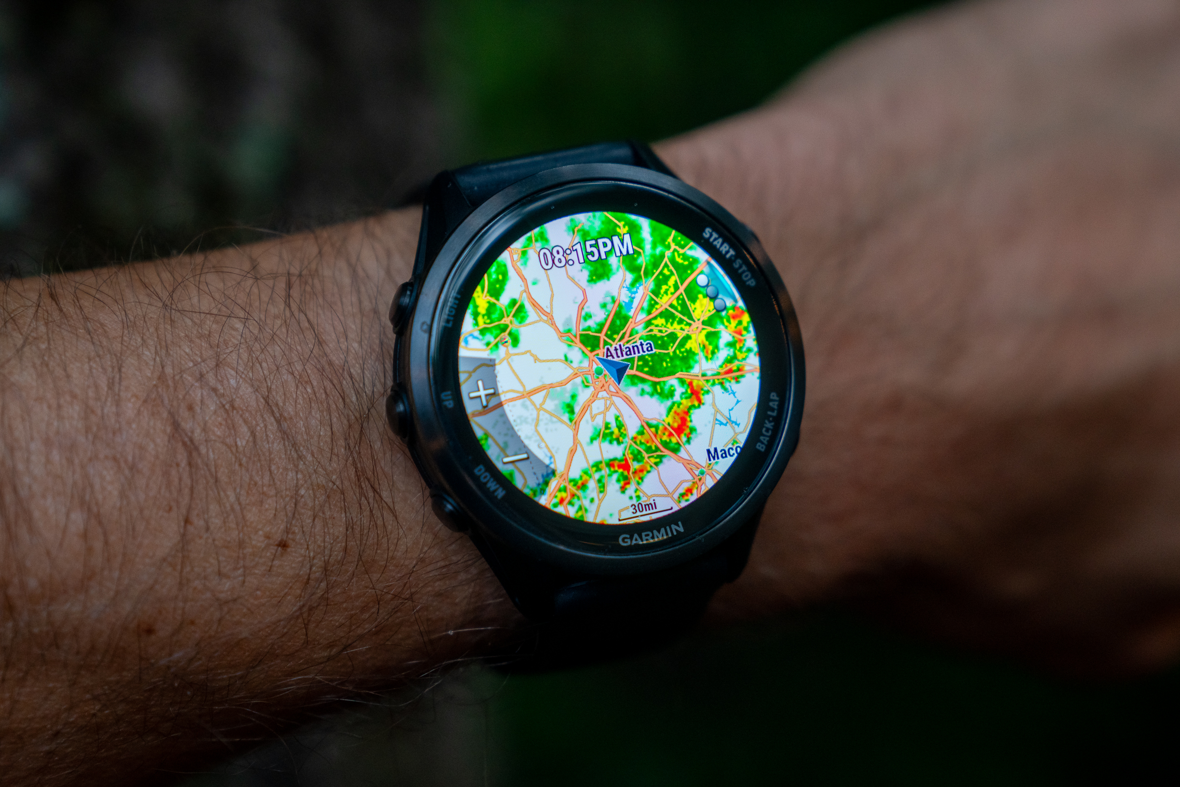 A close-up of a person's wrist wearing a Garmin smartwatch that displays a colorful weather radar map featuring the Atlanta area. The watch screen shows various weather patterns with bright colors indicating precipitation and temperature details. The background is softly blurred, emphasizing the watch.