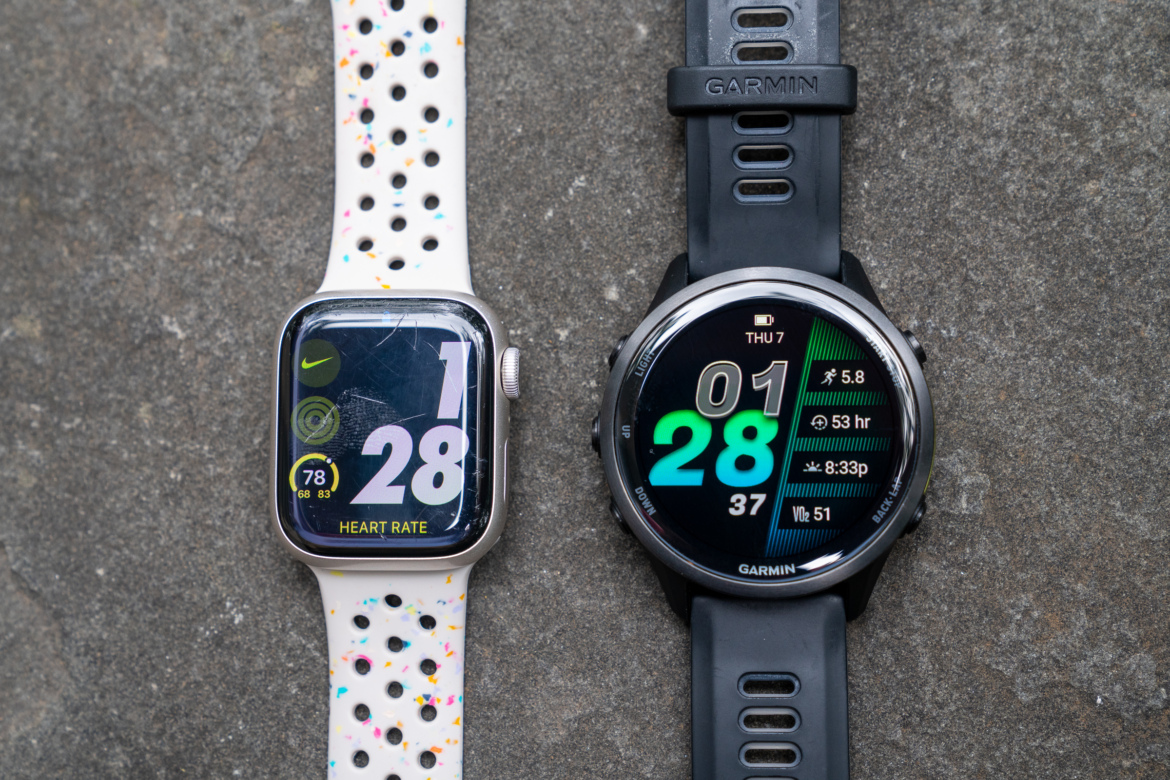 A close-up view of two smartwatches side by side on a stone surface. On the left, an Apple Watch with a white, perforated band displaying a heart rate of 78 bpm and the time 1:28. On the right, a Garmin smartwatch featuring a black strap, showing the time 01:28, alongside various fitness metrics such as a running pace of 5.8 and a VO2 max of 51.
