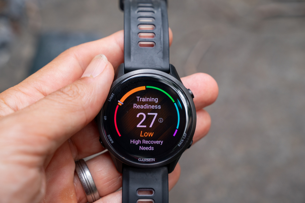 A person holding a black Garmin watch displaying the "Training Readiness" screen, indicating a score of 27, categorized as "Low" with a note about "High Recovery Needs." The background is slightly blurred, focusing on the watch's interface.