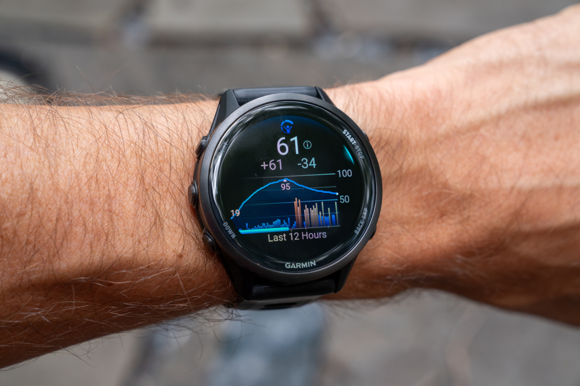 A close-up image of a black Garmin smartwatch on a person's wrist, displaying various health metrics and graphs, including temperature and data for the last 12 hours. The background is blurred, highlighting the watch and the arm.
