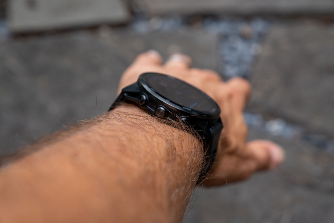 A close-up image of a wrist with a black smartwatch, resting on a stone surface. The watch features a round face and several buttons on the side. The arm is slightly extended, revealing light body hair.