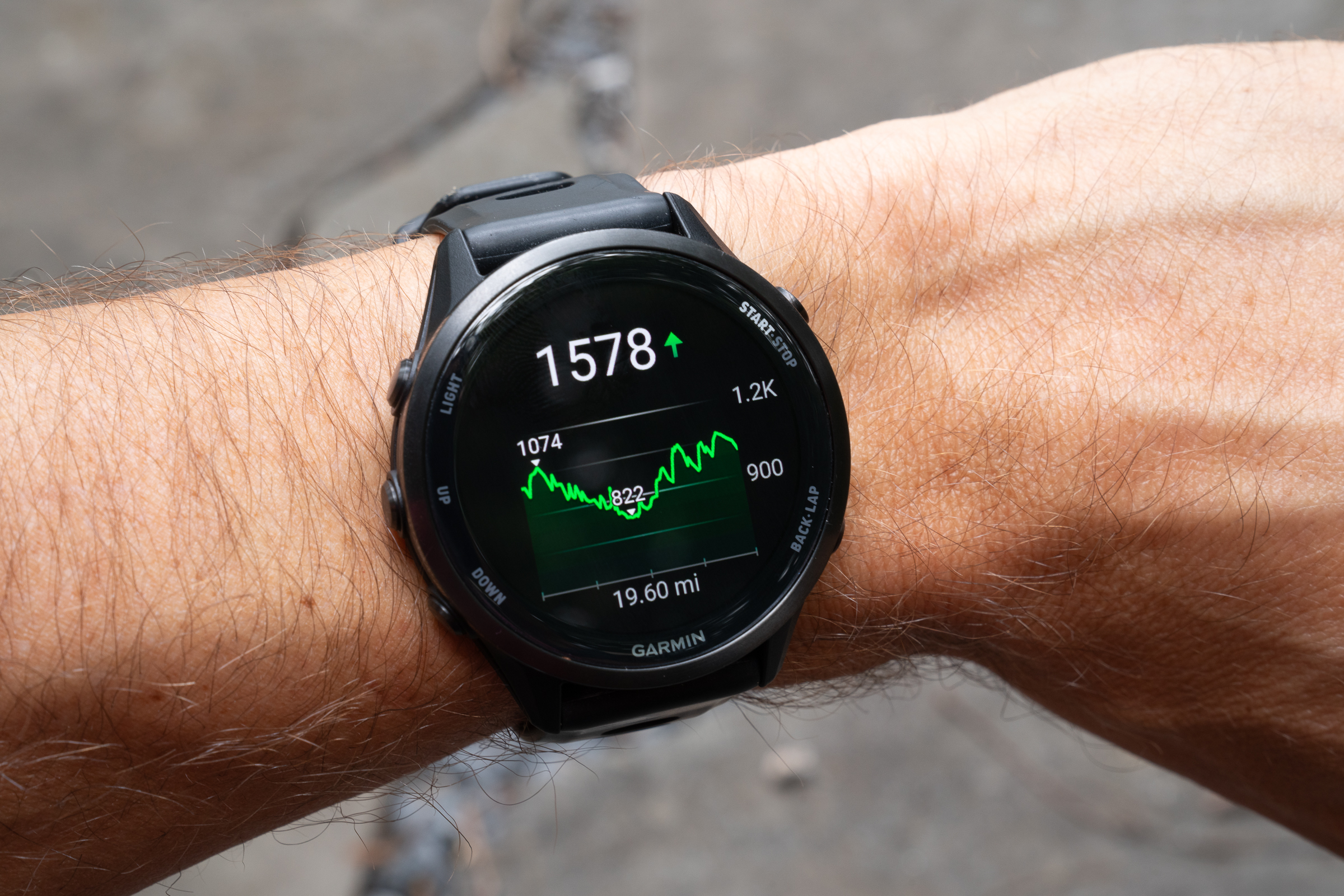A close-up image of a Garmin smartwatch displaying performance metrics, including a heart rate of 1578 BPM and a distance of 19.60 miles, with a green graph line indicating elevation changes. The watch is worn on a person's wrist, showcasing details of the device against a blurred background.