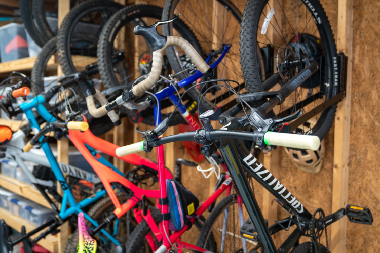 Alt text: A collection of bicycles displayed on a wooden wall, featuring colorful frames in hues of blue, orange, pink, and black. The bicycles are neatly arranged, showcasing their handlebars and tires in a well-organized garage space.