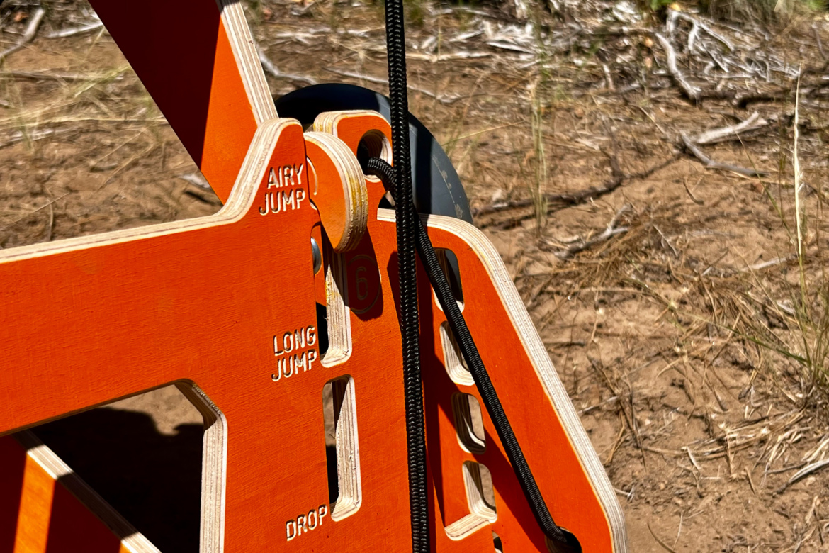Close-up view of an orange wooden training device designed for measuring jumps, with labels for "Airy Jump," "Long Jump," and "Drop." A black cord is attached, and the device is set against a sandy ground with some sparse vegetation in the background.