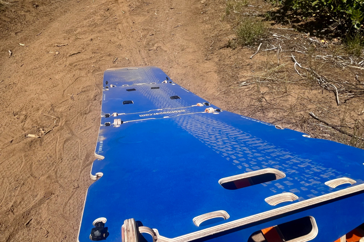 A bright blue rescue board laid flat on a dirt path, surrounded by sparse vegetation and twigs. The board features multiple handles and a textured surface, indicating its use in outdoor rescue operations.