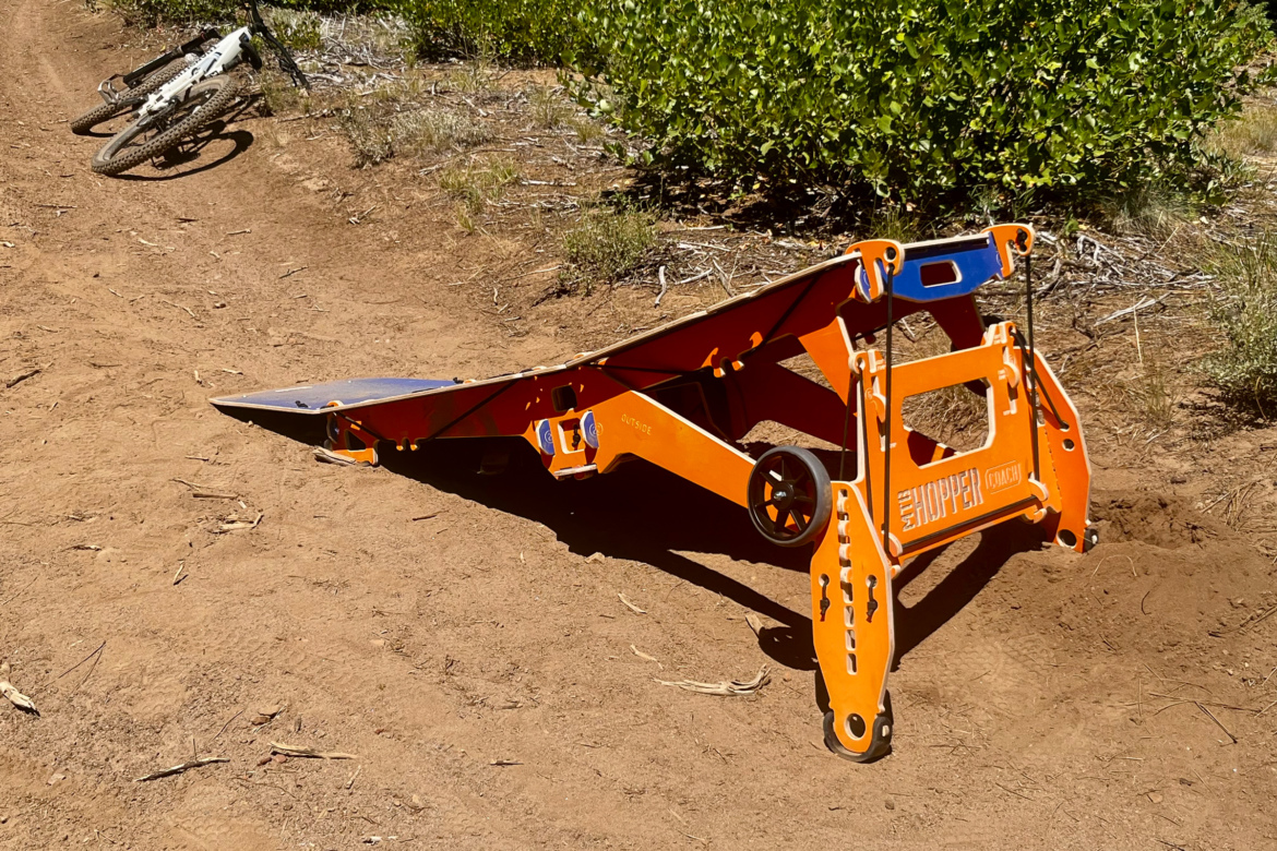 An orange and blue MTB Hopper Coach mountain bike ramp in an outdoor setting, positioned on a dirt trail surrounded by greenery. Nearby, there is a white mountain bike leaning against the trail.