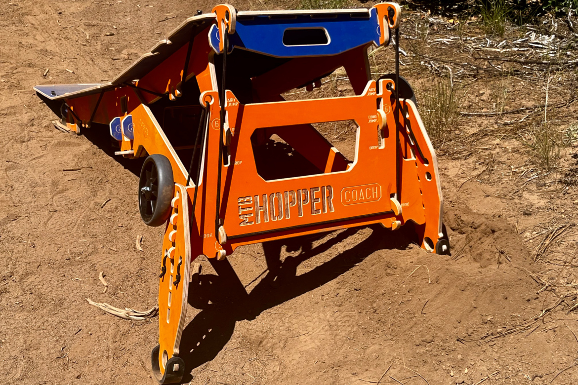 A brightly colored orange and blue MTB Hopper Coach ramp situated on a dirt surface, designed for mountain biking practice and training. The ramp has a collapsible design and features wheels for easy transportation. Sunlight casts shadows on the ground, with some grass and tree debris visible in the background.
