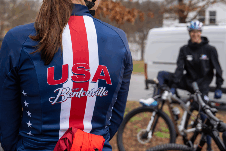 A cyclist wearing a blue jersey with red and white stripes, featuring the text "USA" and "Visit Bentonville," stands in the foreground. In the background, another person in a black jacket and a helmet is preparing a bike. The scene is set outdoors, with some trees in the background and a van parked nearby.