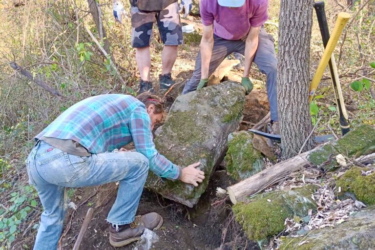Four individuals are working together outdoors, in a wooded area, to lift a large rock. One person is kneeling and pushing against the rock, while another stands nearby, observing. The third person is dressed in a hoodie and shorts, and the fourth is wearing a helmet and holding a tool. The ground is uneven, and there are other rocks and dirt around them, indicating a rugged environment. Trees and foliage surround the scene, suggesting a natural setting. The Local mountain bike trail.
