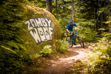 A mountain biker riding along a dirt trail surrounded by lush greenery and trees, with a large boulder on the left side featuring the graffiti text "ZOMBY WOOF." Zombie Woof mountain bike trail.