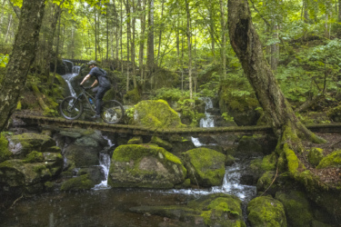 A mountain biker riding over a wooden bridge that crosses a stream, surrounded by lush green foliage and moss-covered rocks. Waterfalls can be seen in the background, enhancing the natural forest setting. Neilson Nord mountain bike trail.