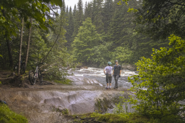 Two mountain bikers stand near a riverbank in a lush, green forest, surrounded by tall trees. One biker is wearing a light blue helmet and has a colorful shirt, while the other is dressed in dark clothing. Their bicycles are positioned nearby, and it appears to be raining lightly, creating a serene atmosphere in nature. Neilson Nord mountain bike trail.