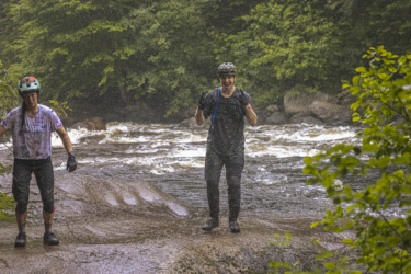 Two individuals stand in a river during rainfall, surrounded by lush green foliage. One person, wearing a colorful helmet and a damp shirt, appears focused, while the other, dressed in a black outfit, smiles and raises a hand in excitement. The rushing water of the river flows behind them, creating a dynamic and adventurous atmosphere. Neilson Nord mountain bike trail.
