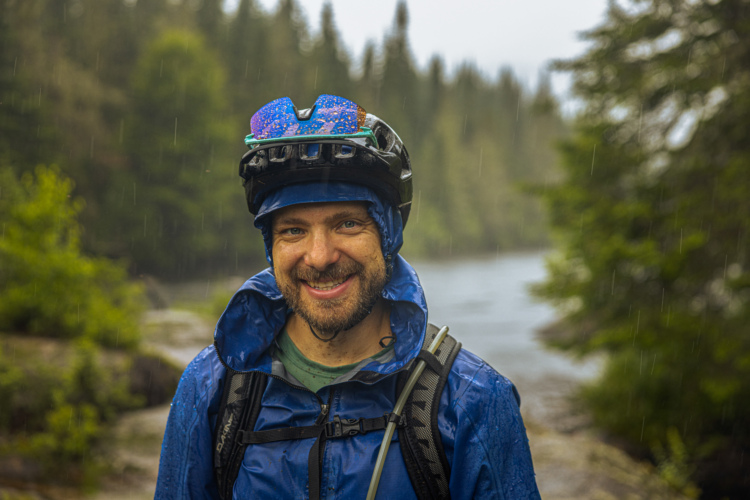 A person wearing a blue rain jacket and a helmet, smiling amidst a rain shower. In the background, a river flows through a lush, green landscape with tall trees. Rain droplets are visible in the air, adding to the outdoor, adventurous atmosphere.