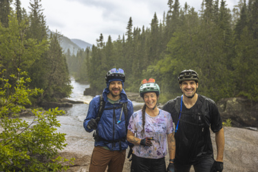 Three mountain bikers smiling and posing for a photo in a lush, green forest near a river, with light rain falling around them. They are wearing helmets and biking gear, surrounded by trees and a scenic backdrop of mountains. Neilson Nord mountain bike trail.