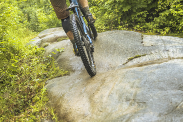 A mountain biker riding up a rocky trail surrounded by lush greenery, showcasing a sense of adventure and outdoor activity. The biker is dressed in a blue jacket and brown shorts, wearing protective gear, and is in motion, navigating the uneven terrain. Neilson Nord mountain bike trail.