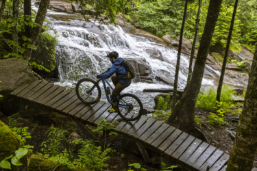 A mountain biker navigating a wooden bridge over a stream, surrounded by lush greenery and a waterfall in the background. The biker is wearing a helmet and blue jacket, showcasing a scenic outdoor adventure in a forested area. Neilson Nord mountain bike trail.