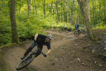A mountain biker leans into a turn on a dirt trail surrounded by lush green trees. In the background, two other cyclists ride along the path, enjoying the natural scenery. The image captures the thrill of outdoor biking in a wooded area. Octopus - Kraken mountain bike trail.