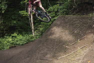 A cyclist in a helmet and a floral-patterned jersey is performing a jump on a dirt bike trail in a forested area. The lush greenery surrounds the scene, providing a vibrant backdrop as the cyclist catches air over a dirt ramp. Octopus - Kraken mountain bike trail.
