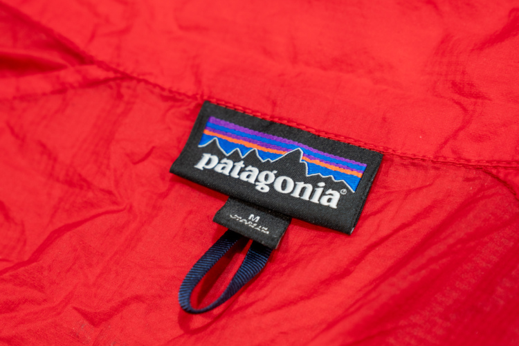 A close-up of a black clothing label featuring the Patagonia logo, set against a vibrant red fabric background. The label displays the brand name "patagonia" along with a mountainous graphic and indicates a size medium.
