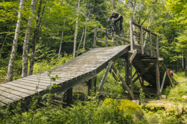 A mountain biker launching off a wooden ramp in a lush forest, with trees and greenery surrounding the trail. Another cyclist can be seen in the background. Le Massif de Charlevoix mountain bike trail.