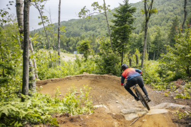 A mountain biker riding a dirt trail through a lush green forest, navigating a steep curve with a backdrop of hills and a distant view of the ocean. Le Massif de Charlevoix mountain bike trail.