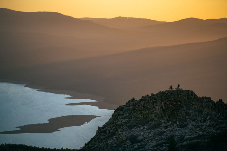 Two mountain bikers are silhouetted against a vibrant sunset, perched atop a rocky outcrop overlooking a winding river and rolling hills in the background. The scene captures a serene landscape with warm golden hues illuminating the sky, transitioning to cooler tones in the distance.