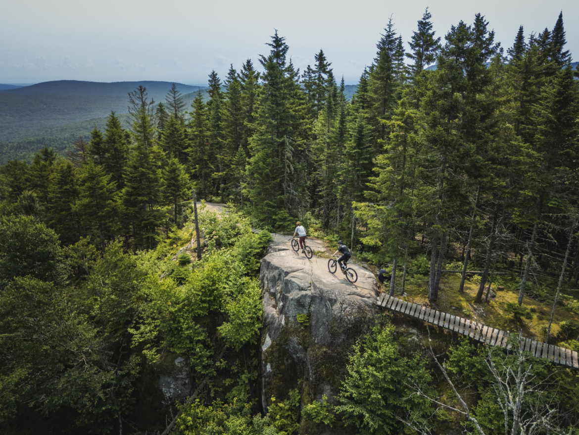 Two mountain bikers navigate a rocky outcrop surrounded by lush green trees, with a scenic view of rolling hills in the background. A wooden bridge extends from the rocky area, leading deeper into the forested landscape. The photo captures the essence of outdoor adventure and the beauty of nature.