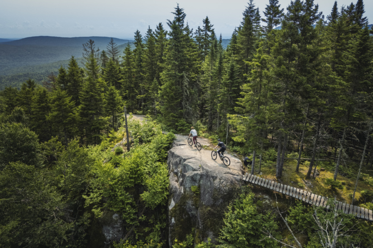 Two mountain bikers navigate a rocky outcrop surrounded by lush green trees, with a scenic view of rolling hills in the background. A wooden bridge extends from the rocky area, leading deeper into the forested landscape. The photo captures the essence of outdoor adventure and the beauty of nature.